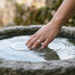 Dłoń zanurzona w przejrzystej wodzie w kamiennej misie, symbol wody jako źródło oczyszczenia i przemiany
