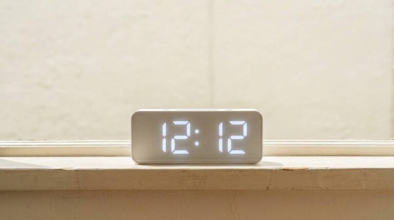 Wyświetlacz cyfrowego zegara pokazujący 12:12 w ostrym świetle południa na jasnym parapecie.